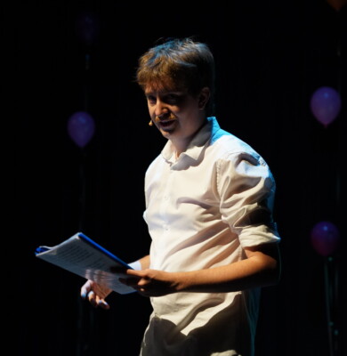Performer Adam Kelly upper body shot. He is with microphone headset, wears white shirt, sleeves rolled up, and holding script.