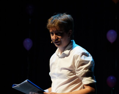 Performer Adam Kelly upper body shot. He is with microphone headset, wears white shirt, sleeves rolled up, and holding script.